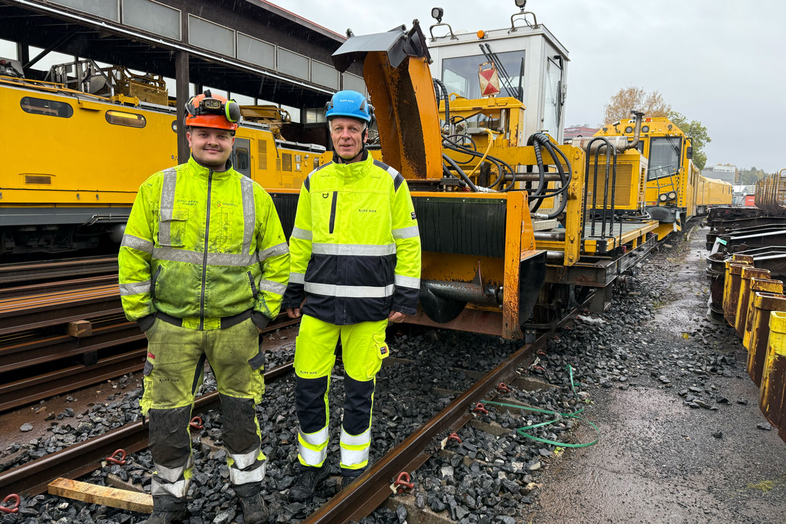 To menn i gule arbeidsklede framfor brøytemaskinar.