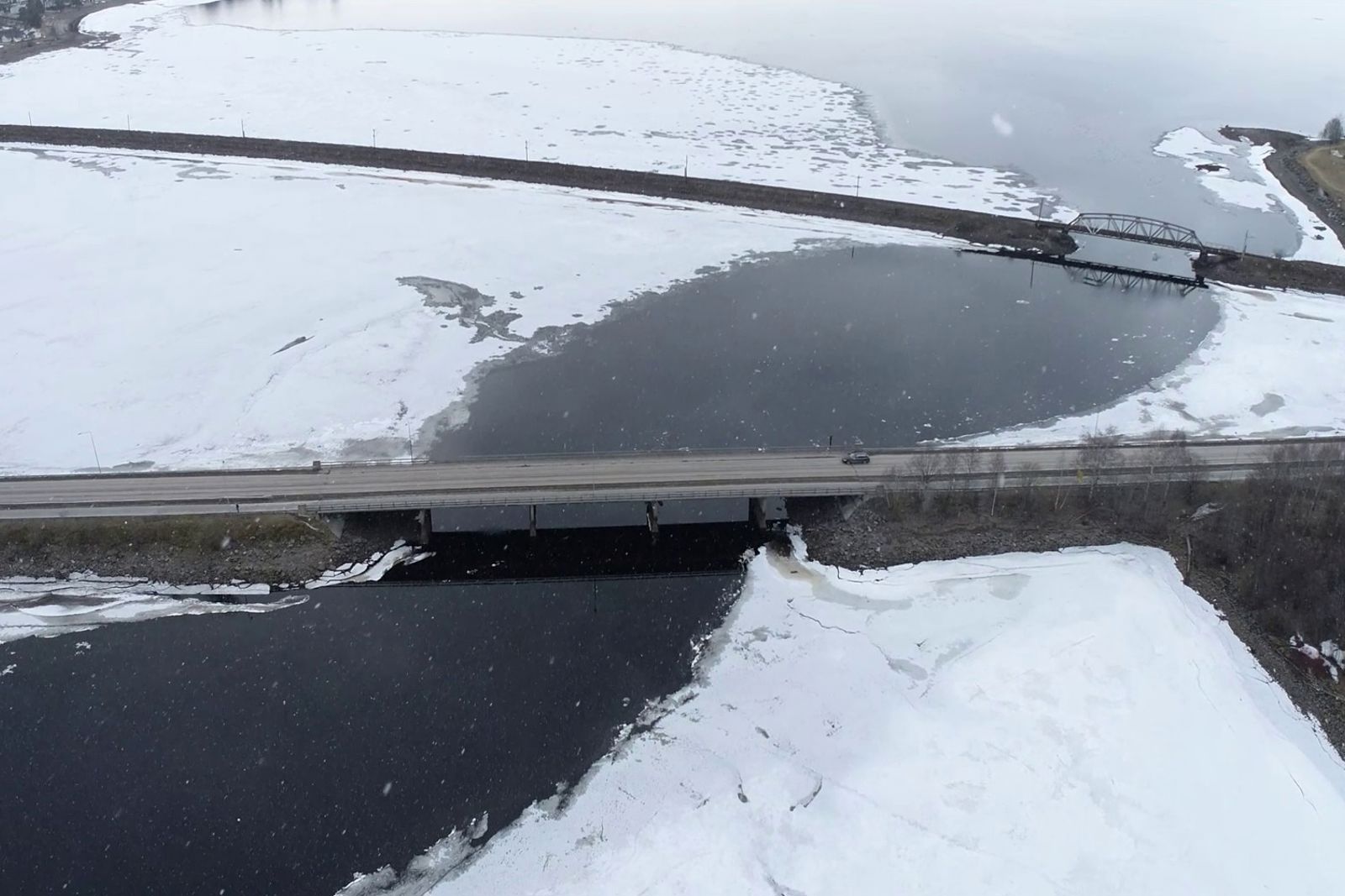 Dronefoto av veg og bane på en kombinasjon av bru og fylling. Snø og is på vannet, med en åpen råk.