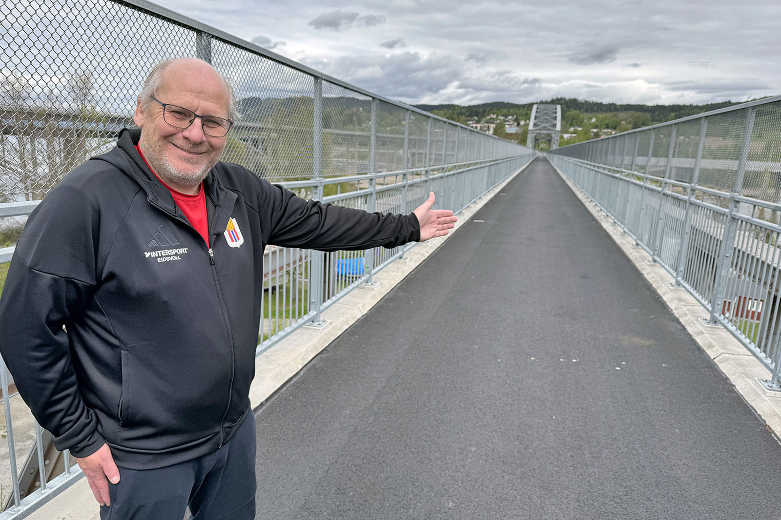 En blid Halberg i treningstøy peker inn på Minne bru, hvor turvegen krysser. Gjerde på begge sider.