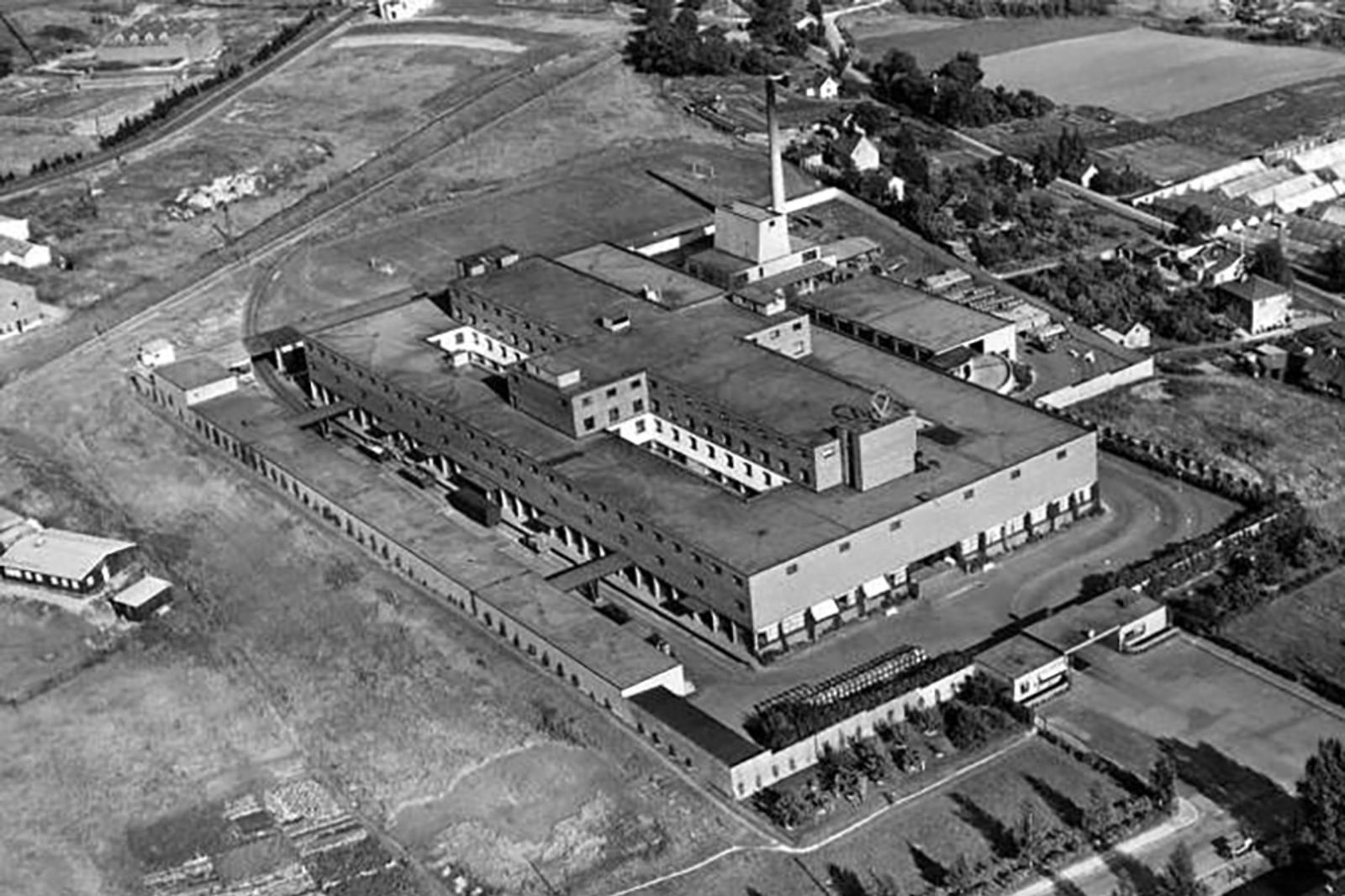Stort industribygg omgitt av jorder og noen villaer.