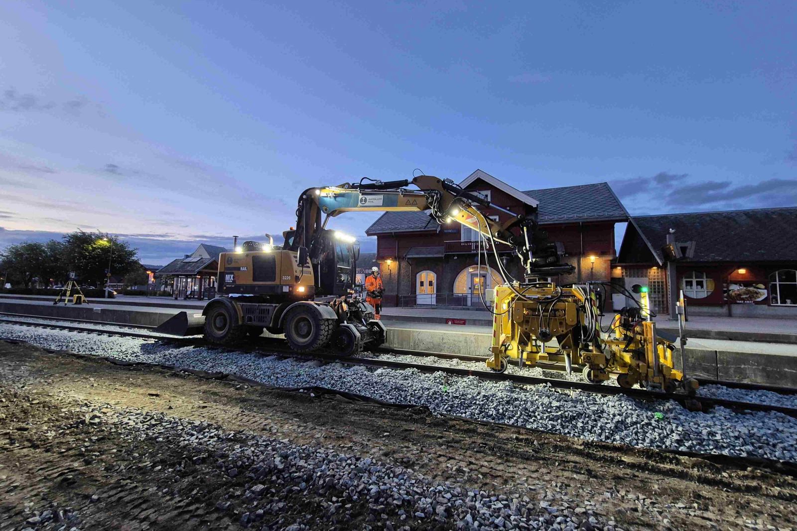 Gravemaskin som jobber med jernbanespor med Verdal stasjon i bakgrunnen