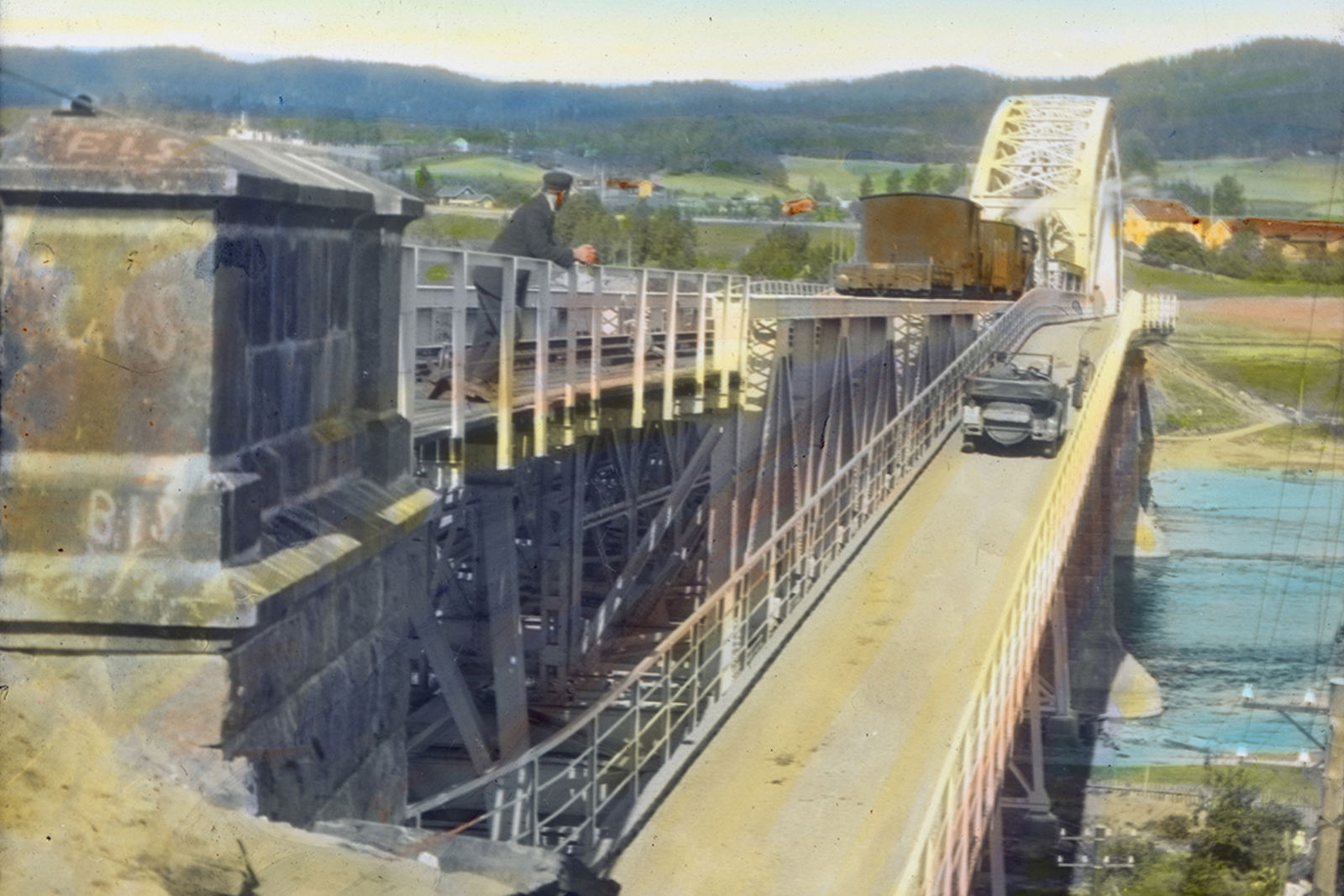 Et tog er oppe på brua og en bil er på veg i kjørebanen. En person er lent mot brurekkverket.